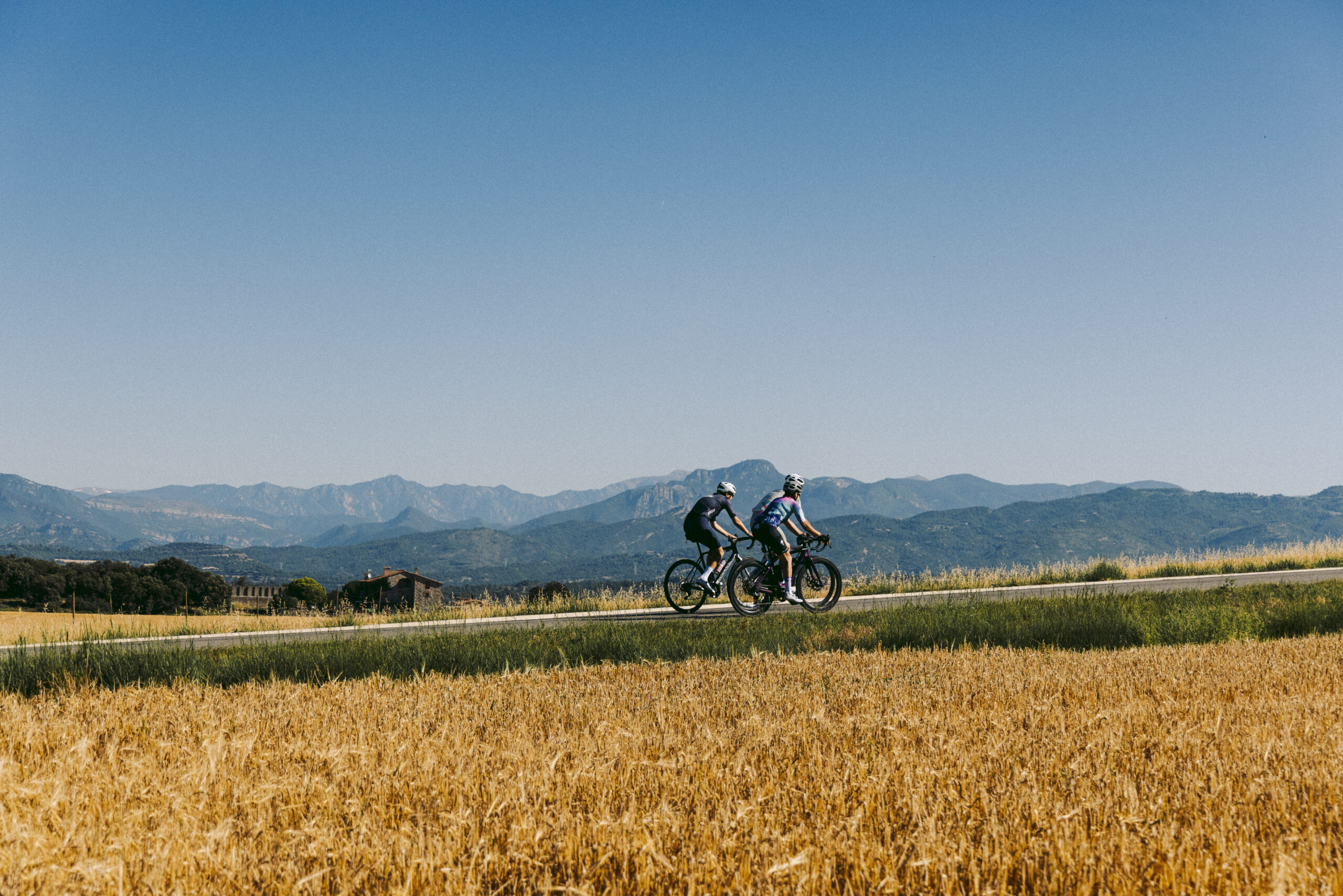 Dos ciclistes en una carretera entre camps amb les muntanyes al fons